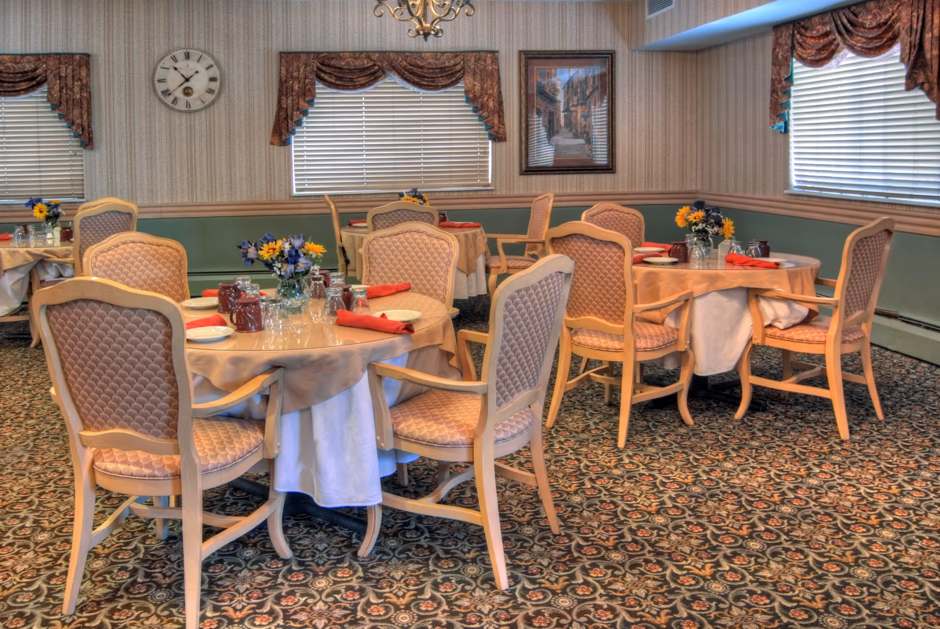 Round dining tables set with plates, glasses, napkins and floral centerpieces surrounded by upholstered chairs in a carpeted dining room with windows and a wall clock.