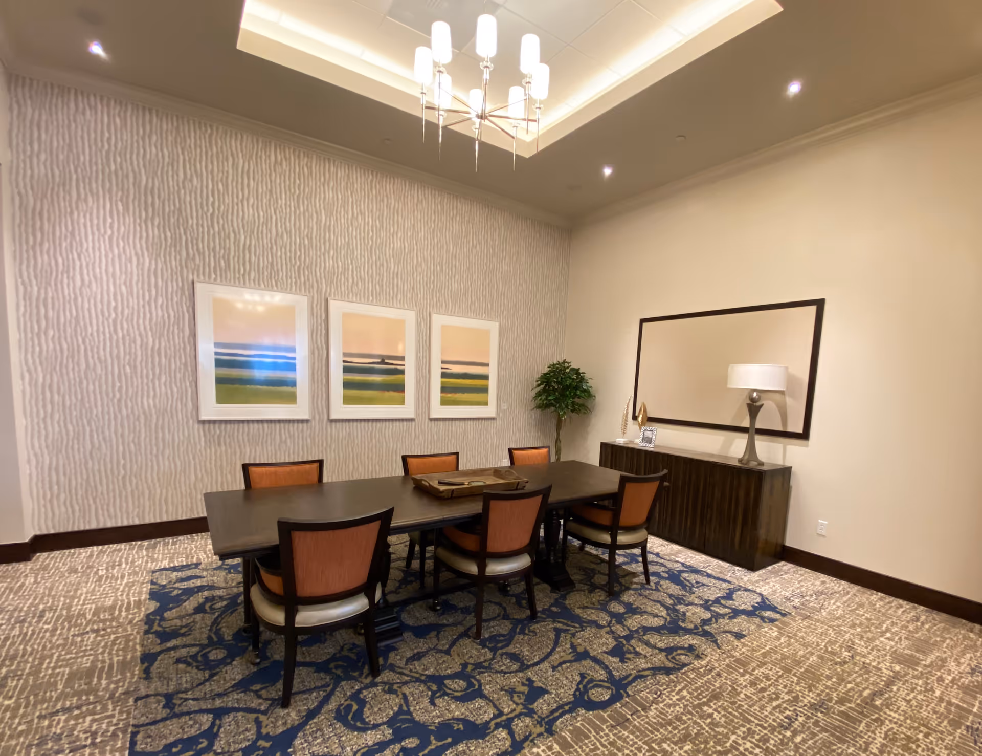 A well-lit meeting/dining room with a long dark table surrounded by six chairs, framed landscape artwork on the wall, a sideboard with a lamp, and patterned carpet.