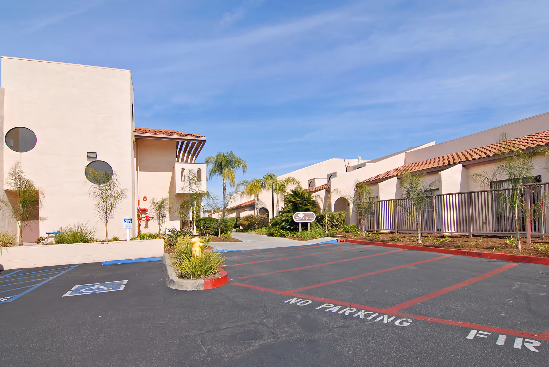 Exterior view of Grossmont Gardens Memory Care facility showing a parking area with marked spaces including a handicapped spot and a no parking fire lane. The building has a light beige stucco finish with red tile roofing, surrounded by palm trees and landscaping under a clear blue sky.
