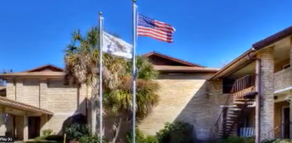 Exterior view of Camelot Chateau Assisted Living facility showing a two-story building with beige brick walls, a staircase on the right side, two flagpoles with an American flag and another flag, palm trees, and clear blue sky.