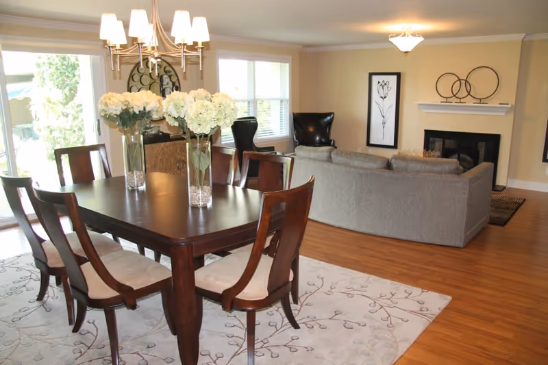 A bright and spacious living and dining area with a dark wooden dining table surrounded by six chairs, three vases with white flowers on the table, a large window letting in natural light, a gray sectional sofa facing a fireplace with decorative items on the mantel, and two black armchairs near the window.
