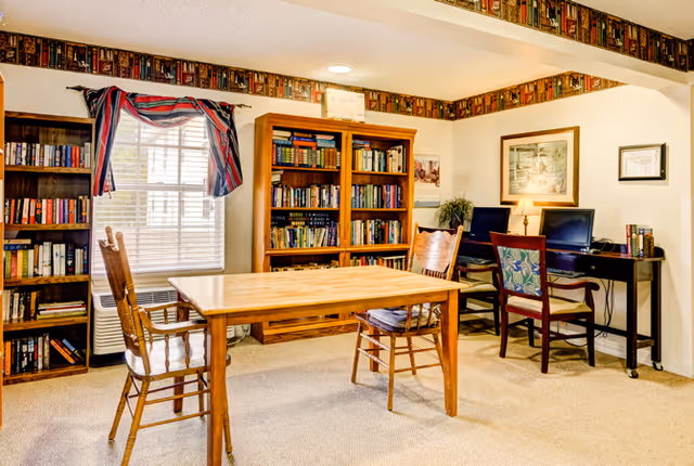 A cozy room with beige carpet featuring a wooden table with two wooden chairs in the center. Behind the table are two tall wooden bookshelves filled with books. A window with blinds and a red, white, and blue valance is on the left wall. On the right side, there is a desk with two computer monitors and two chairs, one with a floral cushion. The walls are decorated with framed pictures and a wallpaper border with a book pattern runs along the top of the walls.