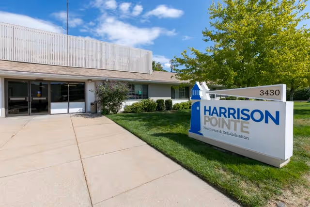 Exterior view of Harrison Pointe Healthcare and Rehabilitation facility showing the entrance with glass doors, a concrete walkway, green lawn, bushes, and a large sign with the facility name and address number 3430 under a partly cloudy blue sky.