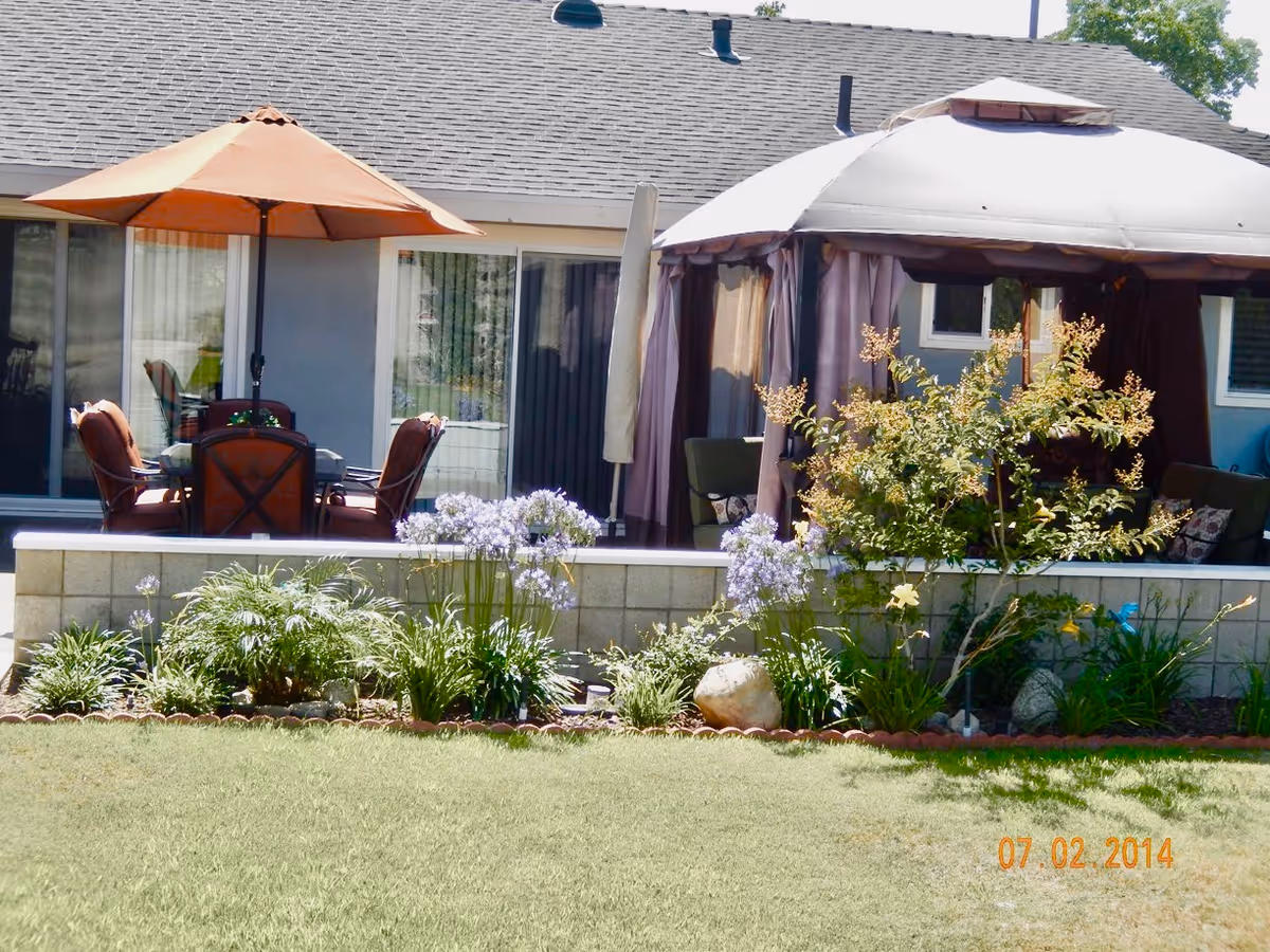 Outdoor patio area with a table and four chairs under an orange umbrella, next to a gazebo with curtains and cushioned seating. There is a garden bed with various plants and flowers in front of a low brick wall, and a grassy lawn in the foreground. The building with sliding glass doors and windows is visible in the background.