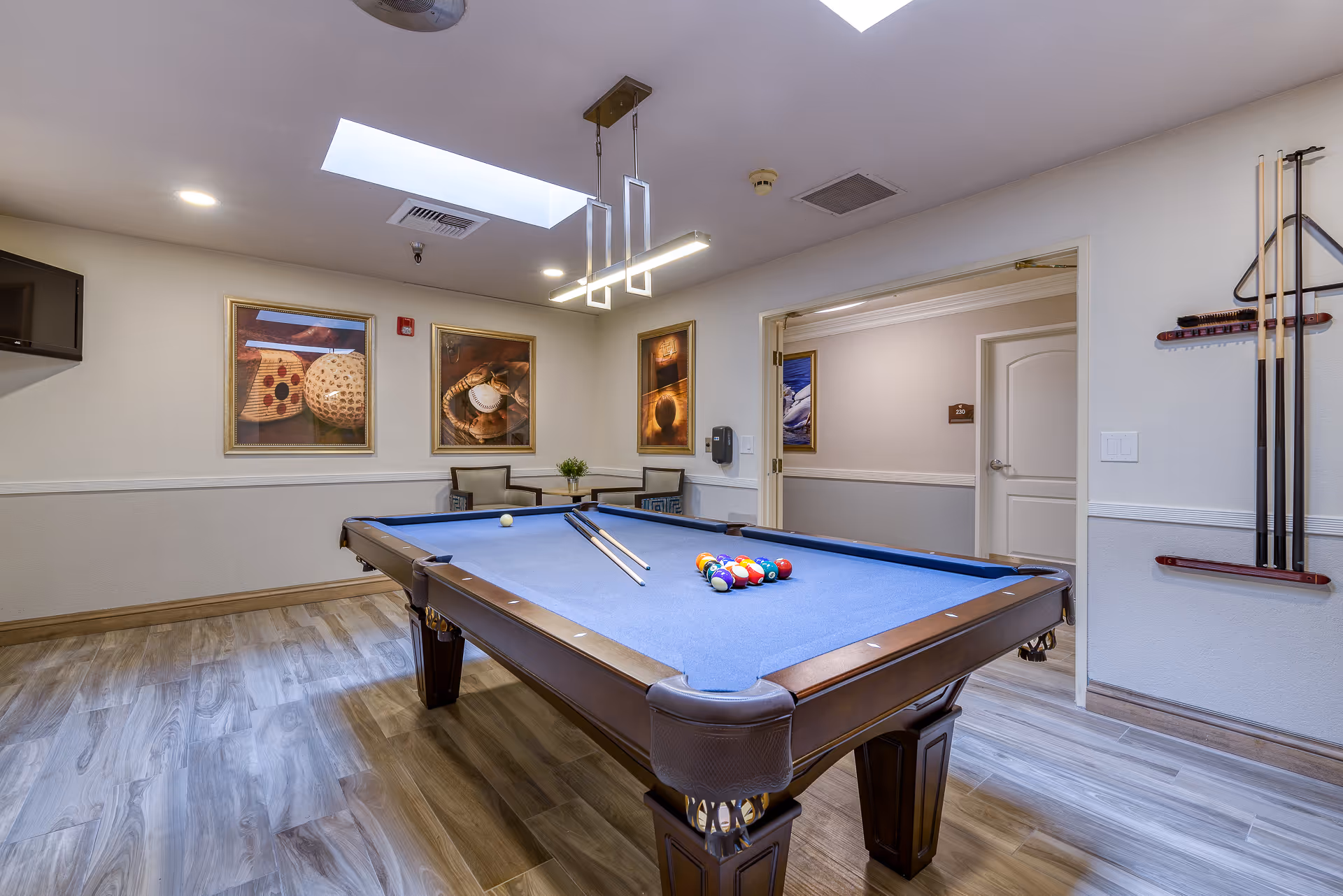 A well-lit game room with a blue felt pool table in the center, two pool cues resting on the table, and a rack of pool cues mounted on the wall. The room has wood-look flooring, three framed pictures on the wall, two chairs with a small table in the corner, a wall-mounted TV, and a skylight in the ceiling.