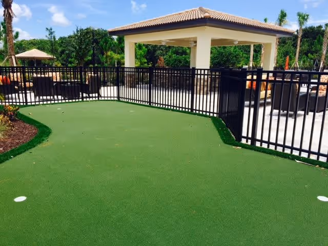 Outdoor area with a putting green surrounded by a black metal fence. In the background, there is a covered pavilion with a tiled roof and outdoor seating including chairs and tables. Palm trees and greenery are visible under a partly cloudy sky.