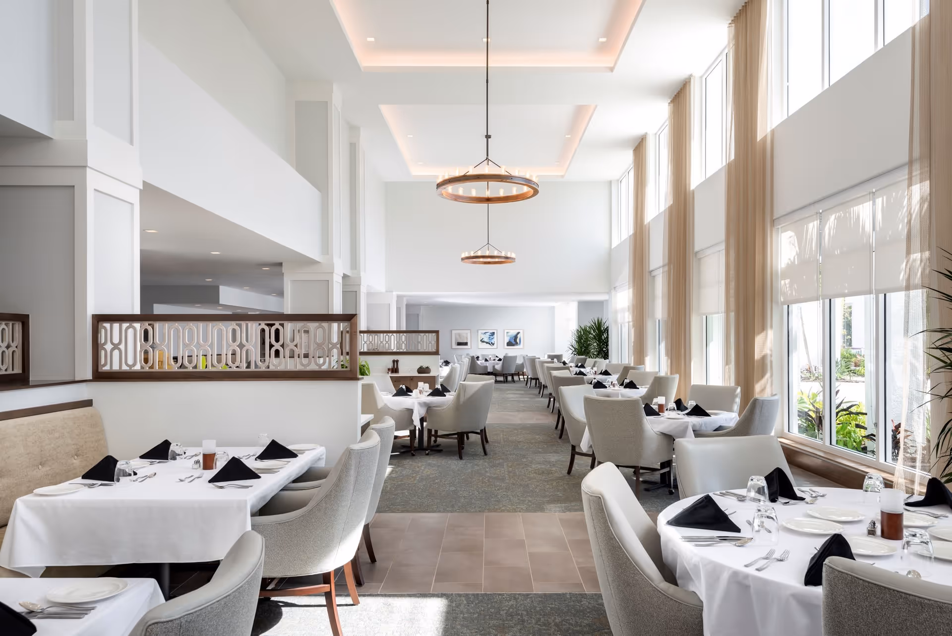 Bright and spacious dining room with high ceilings and large windows letting in natural light. Tables are set with white tablecloths, black napkins, plates, and silverware. Comfortable upholstered chairs surround the tables, and decorative light fixtures hang from the ceiling. Beige curtains frame the windows, and plants add greenery to the space.