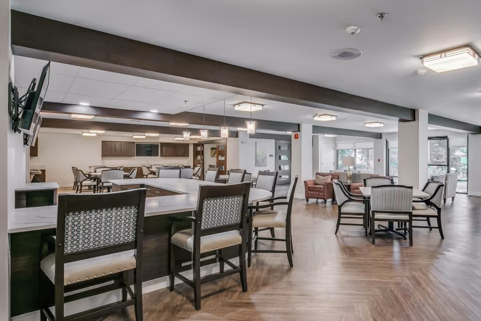 A spacious and well-lit common area in a senior living facility featuring multiple seating arrangements including a large square table with chairs, smaller round tables with chairs, and comfortable armchairs near large windows. The room has wooden flooring, modern light fixtures, and a mounted television on the wall.