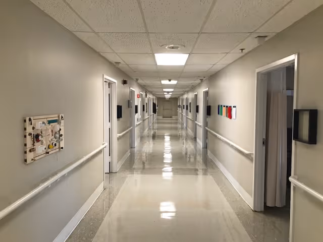 A long, clean, and well-lit hallway in a healthcare facility with beige walls, white handrails on both sides, several open doors leading to rooms, and a shiny tiled floor reflecting the ceiling lights.