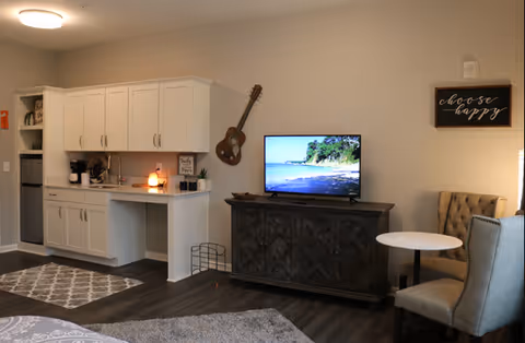 A cozy interior space featuring a kitchenette with white cabinets and a small sink on the left. A guitar is mounted on the wall above a dark wooden TV stand holding a flat-screen TV displaying a beach scene. To the right, there is a small round table with two upholstered chairs. A framed sign on the wall reads 'choose happy'. The floor is covered with dark wood and area rugs.