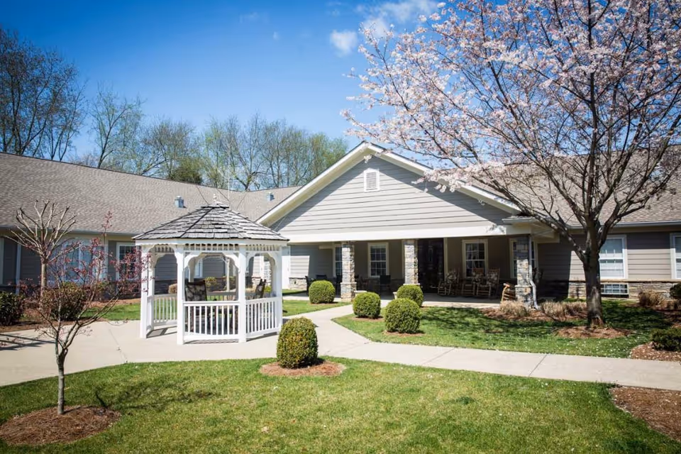 Outdoor courtyard area of Chandler Park Assisted Living featuring a white gazebo, manicured bushes, a blossoming tree, and a building with a covered porch and rocking chairs under a clear blue sky.