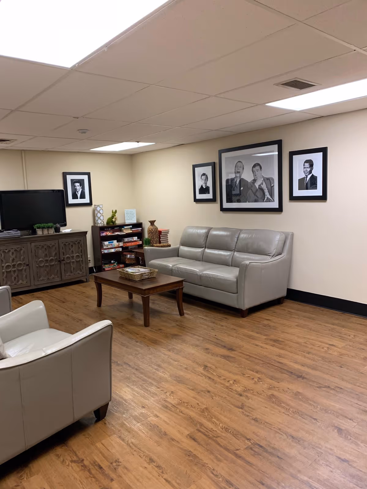 A cozy common room with gray leather sofas, a coffee table, TV cabinet, bookshelves and framed black-and-white portraits on the wall.