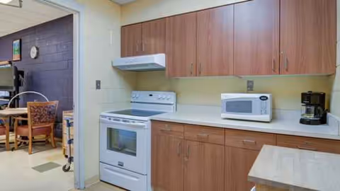 A communal kitchen area with wooden cabinets, stove, microwave, coffee maker, and adjacent dining area.