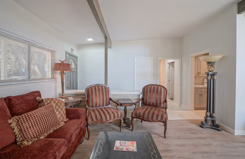 A cozy living room with a red sofa adorned with patterned cushions, two striped armchairs with wooden frames, a small wooden side table between the chairs, a tall decorative floor lamp, and a window with blinds. The room has light-colored walls and wood flooring, with a view into an adjacent kitchen area.