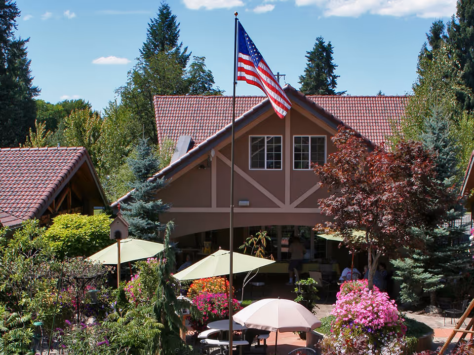 Outdoor patio area at Courtyard at Coeur d'Alene featuring a building with a red-tiled roof, an American flag on a flagpole, several green umbrellas shading tables, and lush greenery including trees and flowering plants.