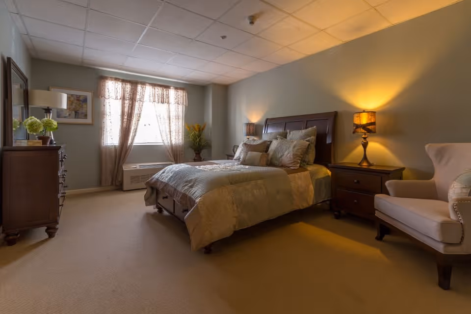 A cozy bedroom with a large bed covered in a beige and green quilt, multiple pillows, two wooden nightstands each with a lamp, a beige armchair, a wooden dresser with a lamp and a vase of flowers, a window with sheer curtains, and a framed picture on the wall.