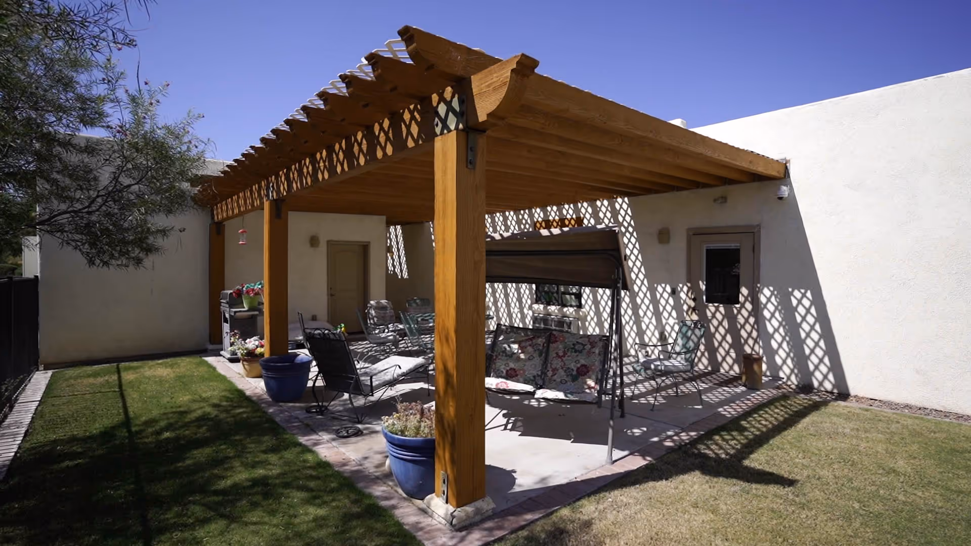 Outdoor patio area with a wooden pergola casting lattice shadows on the ground and walls. The patio is furnished with metal chairs, a swing bench with floral cushions, and potted plants. The surrounding area has grass and a tree on the left side.