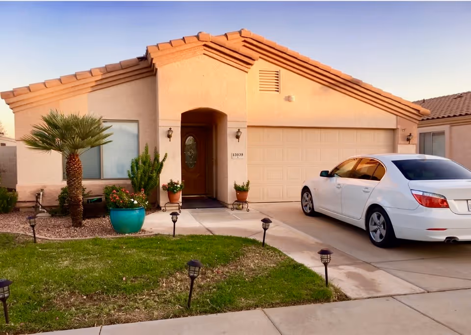 Front exterior view of a single-story house with a beige stucco finish and a tiled roof. There is a driveway with a white sedan parked on it, a small lawn with garden lights, and several potted plants near the entrance door.