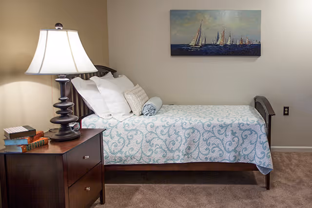 A neatly made single bed with a blue and white patterned bedspread, several pillows, and a bolster pillow. Next to the bed is a dark wooden nightstand with a lamp and a few stacked books. A painting of sailboats on the water hangs on the beige wall above the bed.