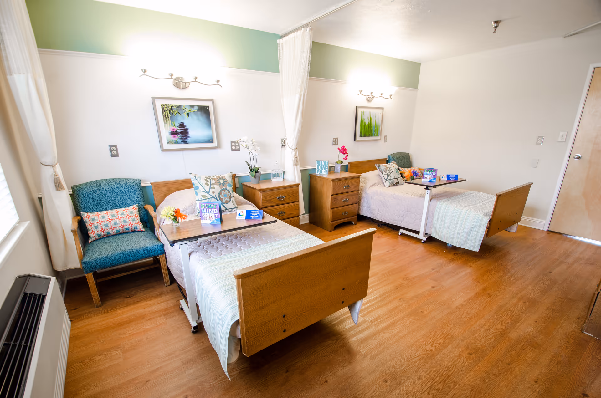 Bright senior facility room with two twin beds, bedside tables, chairs, and framed wall art.
