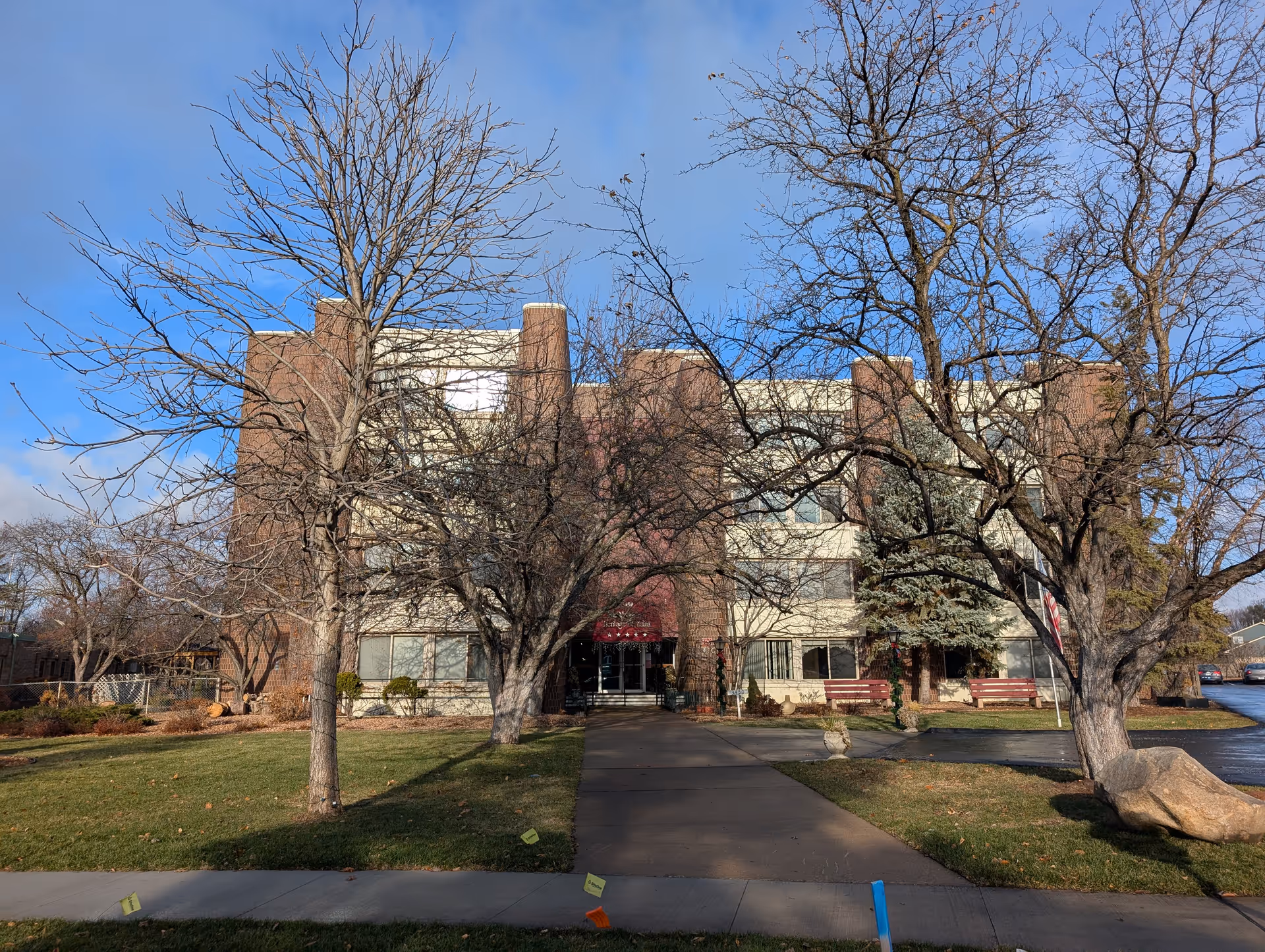 Front exterior view of a multi-story senior living facility building with a paved walkway leading to the entrance. Leafless trees and green grass are visible in the foreground under a partly cloudy blue sky.
