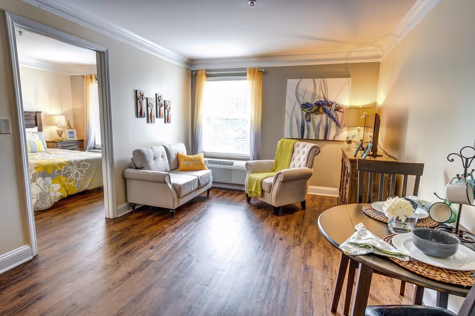 A bright and cozy living area with wooden flooring, featuring a small loveseat with a yellow pillow, an armchair with a green throw blanket, a wooden sideboard with a lamp and decorative items, and a round dining table set with plates, bowls, and napkins. A window with yellow curtains lets in natural light. In the background, a bedroom with a bed covered in a yellow and gray floral comforter is visible through an open doorway.