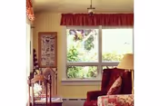 A cozy living room with a large window showing greenery outside. The room features a red upholstered armchair, a floral-patterned sofa, a wooden chair with a woven seat, a floor lamp with a beige shade, and a framed picture on the wall. The window has a red valance curtain.