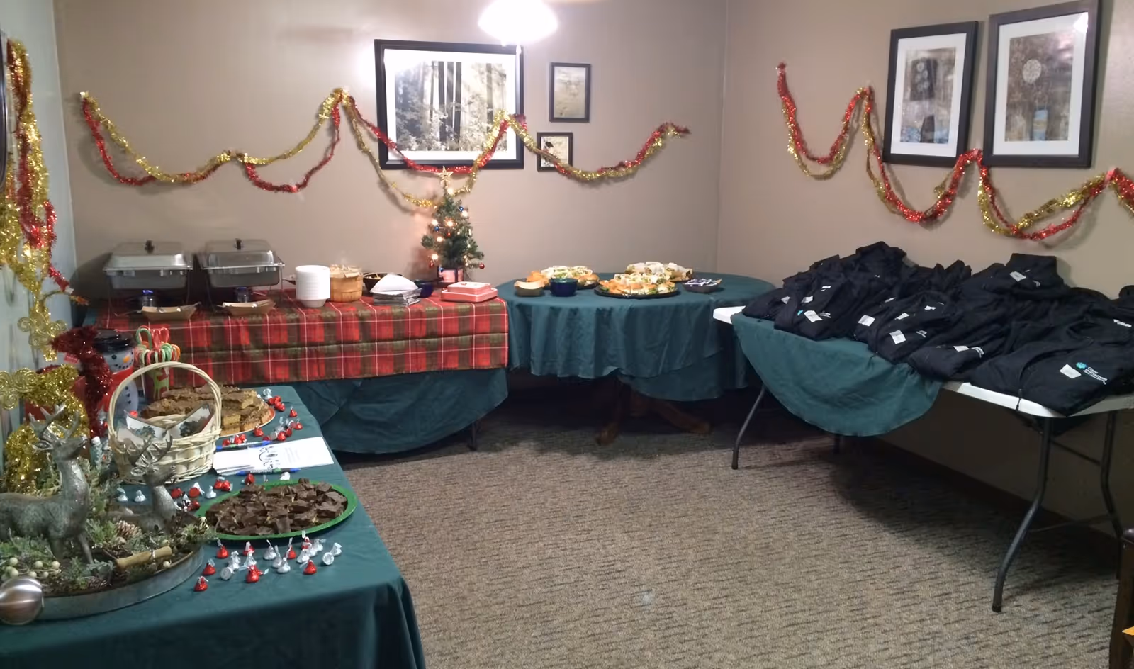 A room decorated for a holiday event with red and gold tinsel garlands on the walls. Several tables covered with green and red plaid tablecloths display trays of food, snacks, and a small decorated Christmas tree. One table holds stacks of black folded clothing items. The walls have framed pictures hanging above the tables.