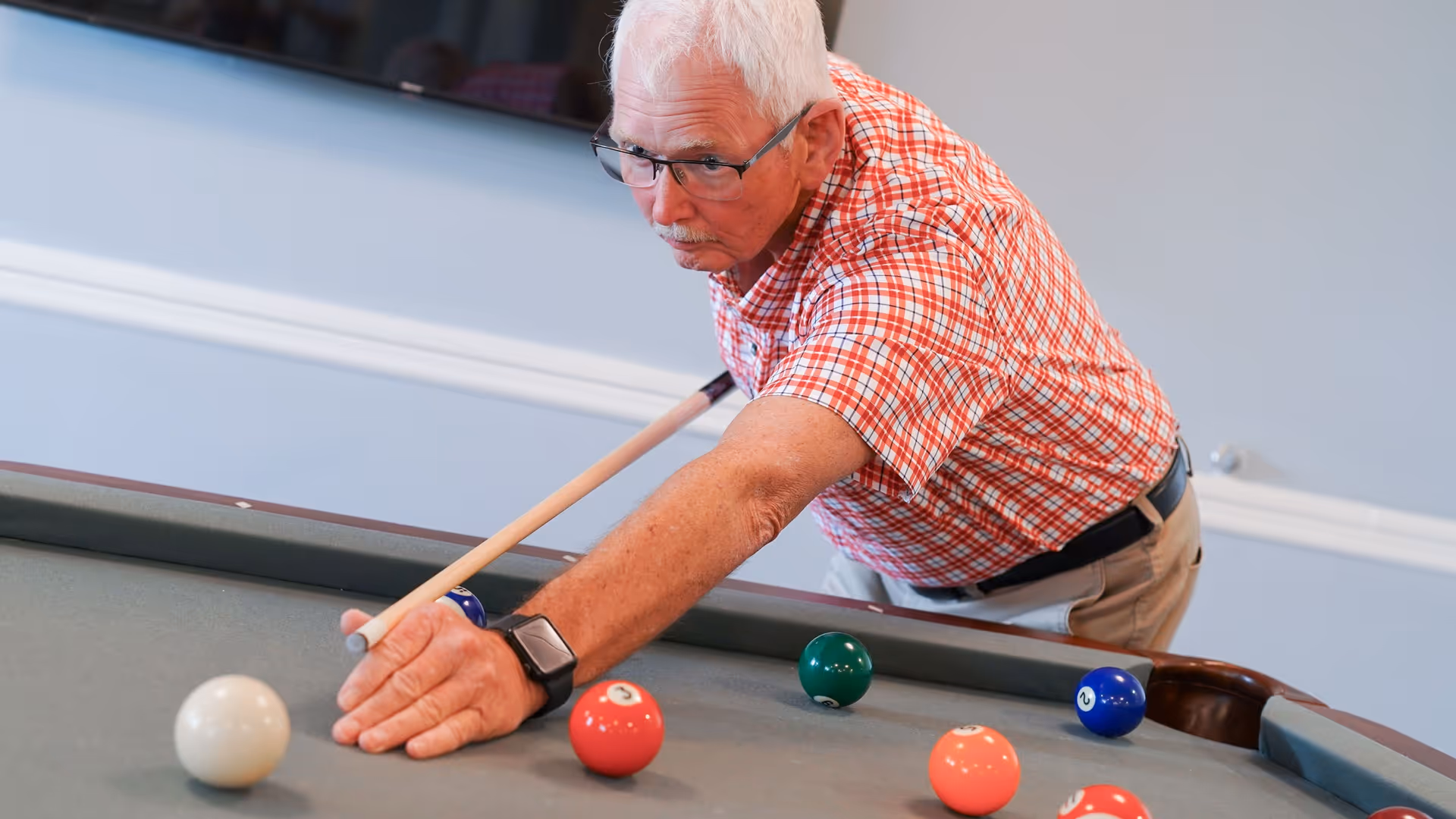 An elderly man wearing glasses and a red plaid shirt is playing pool indoors, aiming with a cue stick at a billiard table with several colored balls visible.
