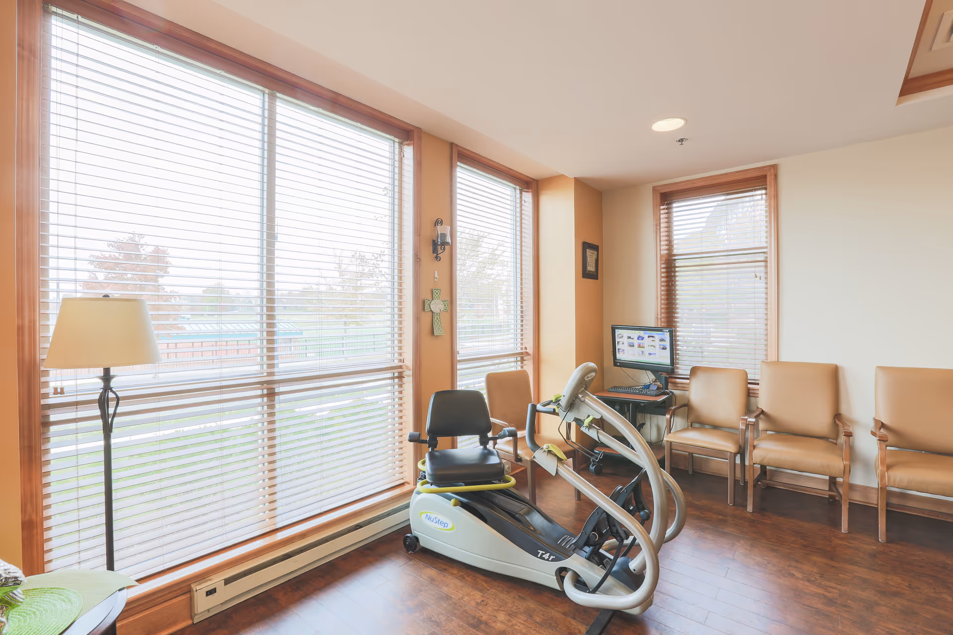 A bright room with large windows covered by blinds, featuring a NuStep exercise machine in the center. There are four tan chairs lined up against the wall, a floor lamp near the window, and a computer on a small table in the corner.