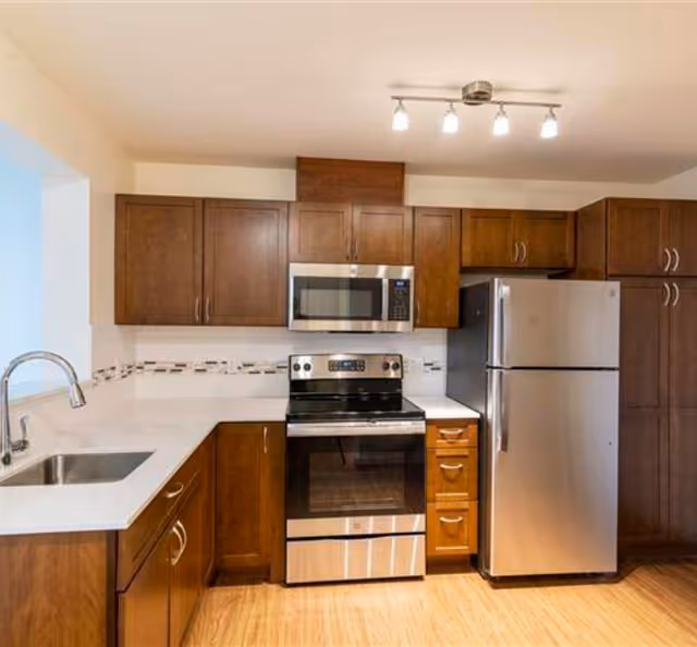 Modern kitchen with wooden cabinets, a stainless steel refrigerator, oven, microwave, and a sink with a chrome faucet. The countertop is white, and there is a decorative tile backsplash. The floor is wooden, and a ceiling light fixture with four bulbs is visible.