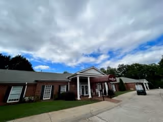 Exterior view of a single-story brick building with a covered entrance and a maroon awning. The sky is partly cloudy with patches of blue visible. There is a driveway and some greenery around the building.