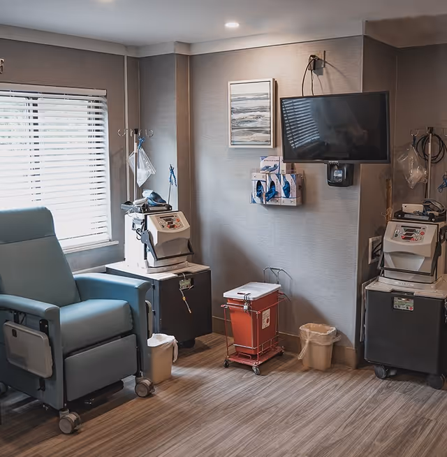 A medical treatment room with a blue reclining chair, two medical machines on wheeled stands, a red biohazard disposal container, a wall-mounted TV, and a window with blinds letting in natural light.