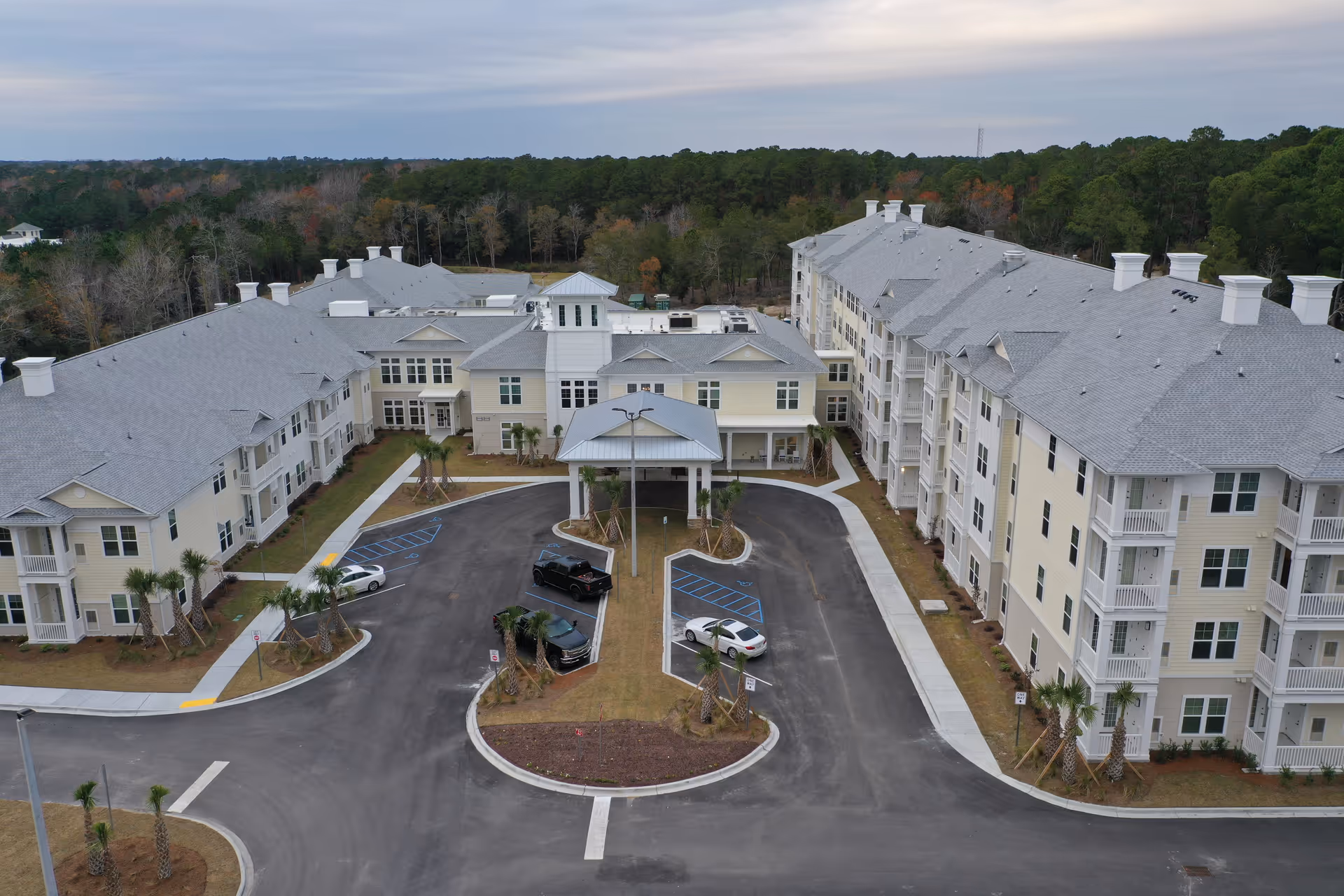 Aerial view of a large senior living facility with multiple connected buildings, a covered entrance, parking spaces including handicapped spots, surrounded by trees and greenery under a cloudy sky.