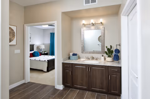 Interior view of a living space in Canyon Winds Independent Living showing a vanity area with a granite countertop, dark wood cabinets, a decorative mirror, and a light fixture above. To the left, there is an open doorway leading to a bedroom with a bed, nightstand, lamp, and blue curtains.