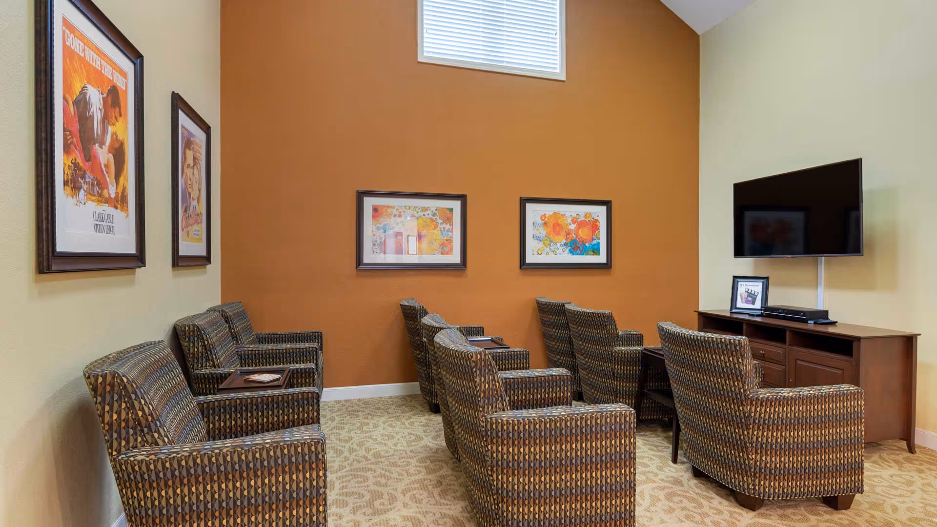 Seating area with several patterned armchairs arranged in rows facing a wall-mounted TV, with framed artwork on orange and beige walls.