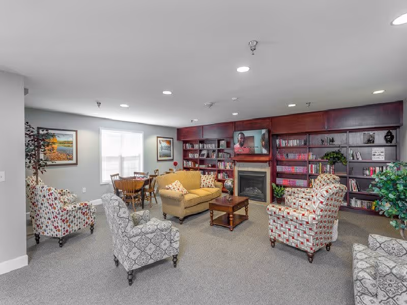 A cozy living room area in a retirement community featuring a yellow loveseat, four patterned armchairs, a wooden coffee table, and a built-in dark wood bookshelf with a fireplace and a TV mounted above it. There is a small round dining table with chairs near a window, and framed artwork on the walls. The room is carpeted and well-lit with recessed ceiling lights.