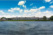 Wide view of a large multi-story retirement living facility named Sandhill Cove Retirement Living, situated along the edge of a body of water under a partly cloudy blue sky.