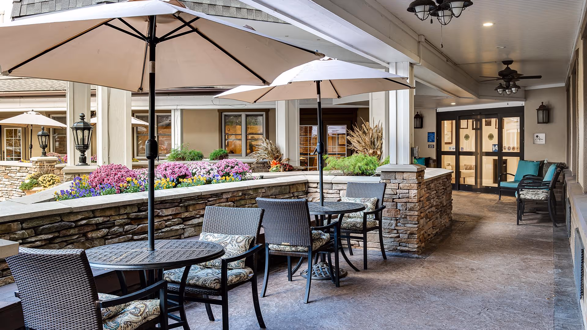 Outdoor covered patio area with round tables and wicker chairs with cushions, large beige umbrellas providing shade, stone half-walls with colorful flowers planted on top, ceiling fans and light fixtures overhead, and double glass doors leading inside the building.
