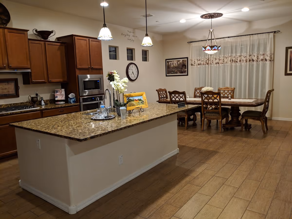 Interior view of a kitchen and dining area in Azalea Assisted Living. The kitchen features wooden cabinets, a granite countertop island with a sink, and hanging pendant lights. The dining area has a wooden table with six chairs, a decorative chandelier, and sheer curtains on the window. The walls are decorated with framed artwork and a clock.