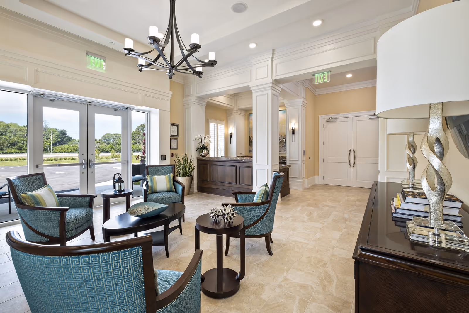 A bright and elegant seating area in a senior living facility lobby with four teal upholstered chairs arranged around two dark wood coffee tables. The room features large glass doors leading outside, beige tiled floors, cream-colored walls with white trim, a modern chandelier, and a dark wood reception desk in the background. A console table with decorative lamps and books is visible on the right side.