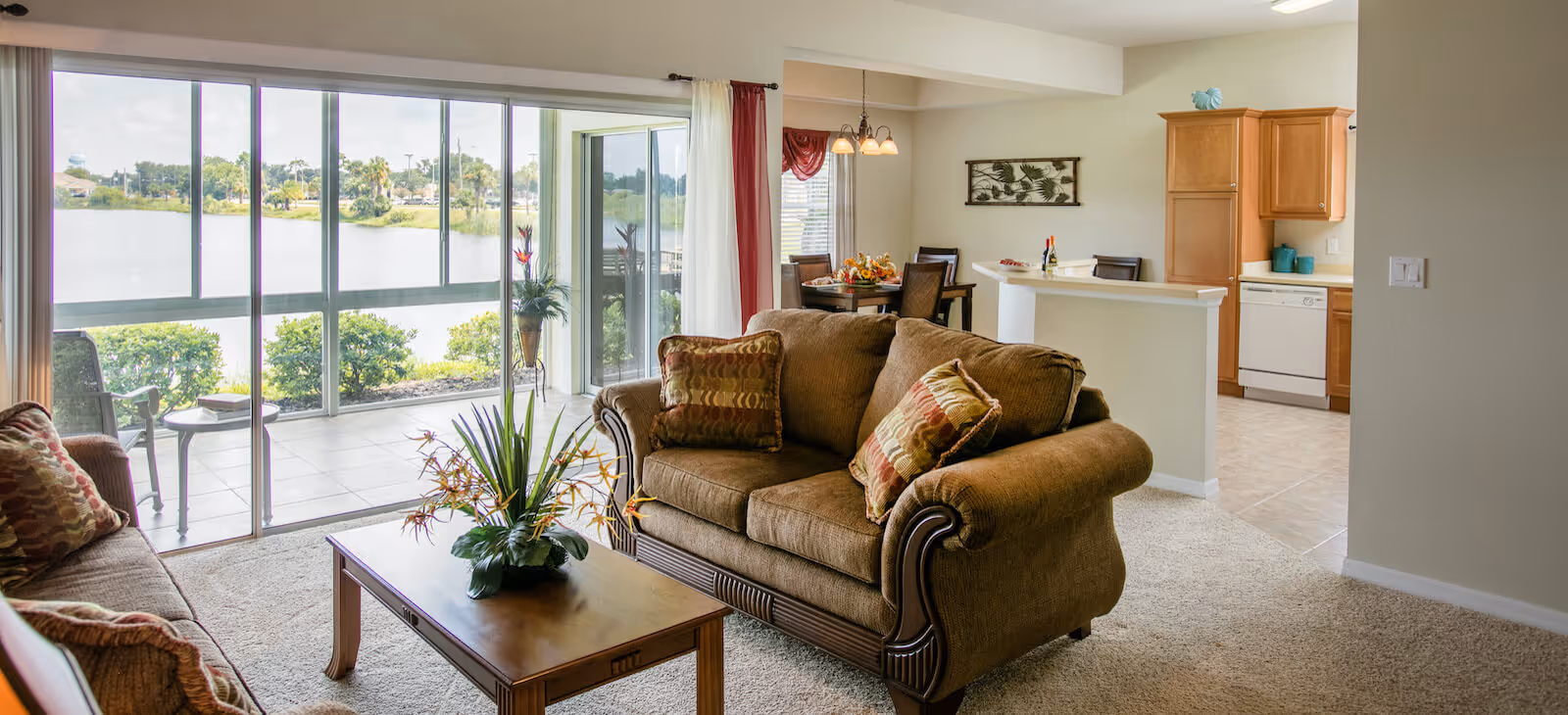 A cozy living room with two brown upholstered sofas adorned with patterned cushions, a wooden coffee table with a floral arrangement, and large sliding glass doors that open to a balcony overlooking a lake. In the background, there is a dining area with a table and chairs, and a kitchen with wooden cabinets and white appliances.