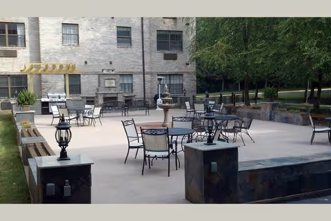 Outdoor patio area with multiple metal tables and chairs arranged on a concrete surface. There are decorative stone pillars with lantern-style lights, a built-in grill, and a stone fireplace against a light brick building. Trees and greenery are visible in the background.