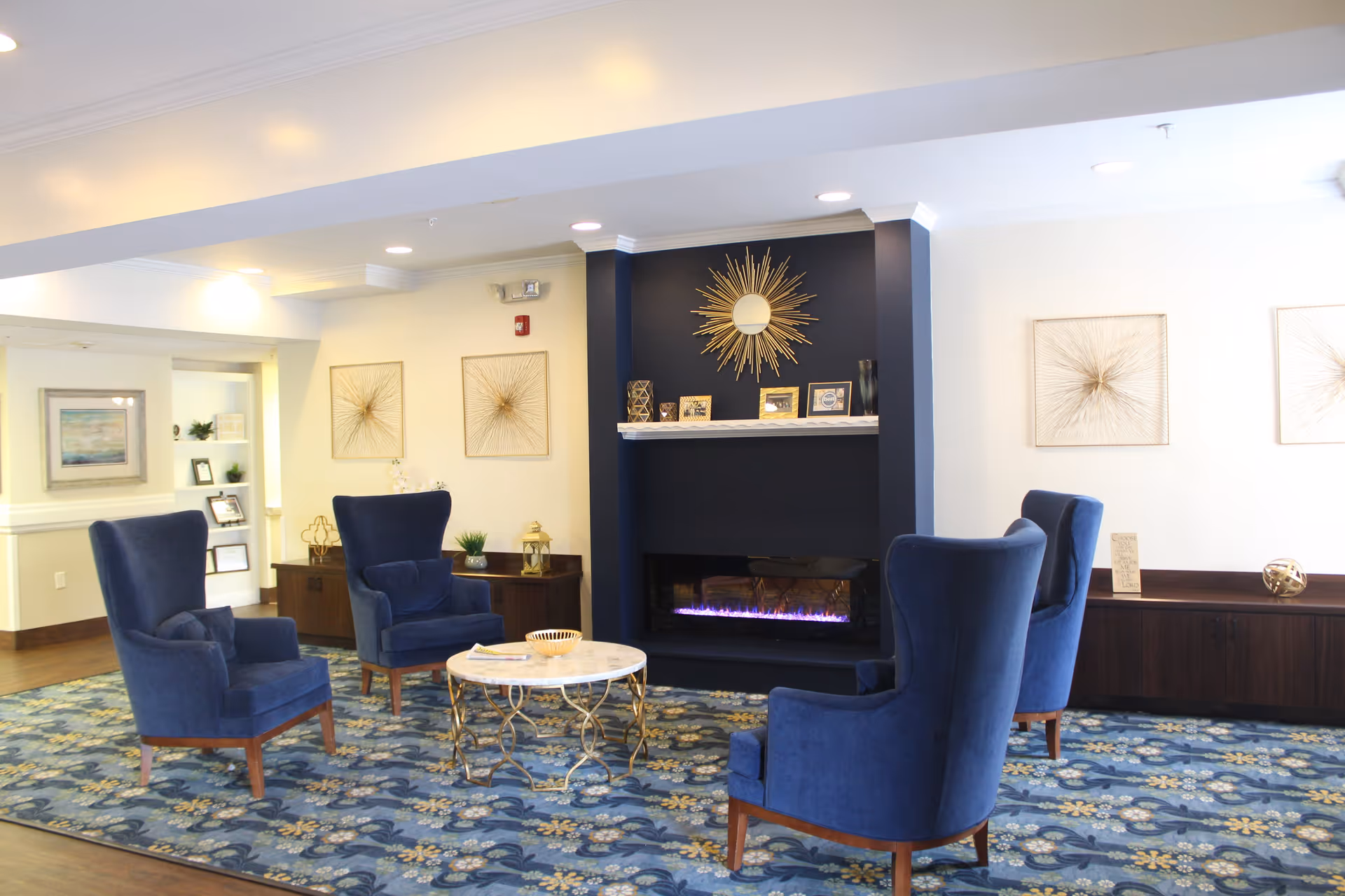 A cozy living room area with four blue upholstered armchairs arranged around a round marble-top coffee table. The room features a dark blue fireplace with a modern electric fire and a decorative sunburst mirror above it. The walls are adorned with abstract gold artwork, and the floor has a blue floral patterned carpet. The space is well-lit with recessed ceiling lights.
