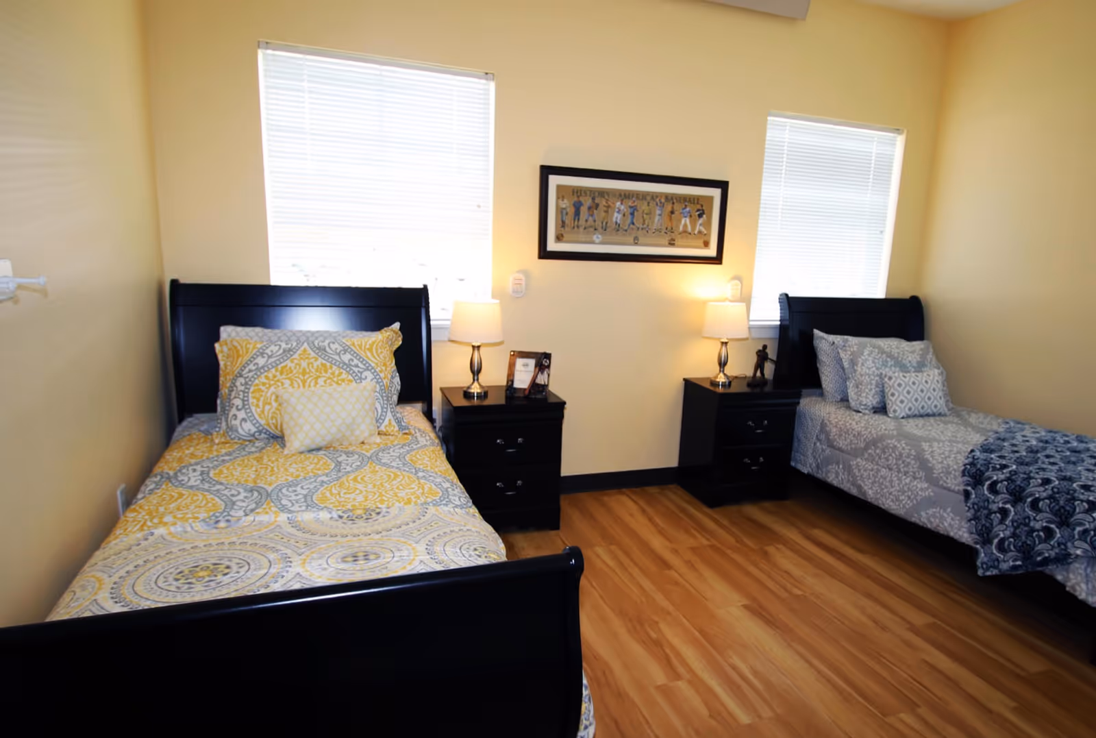 A bedroom with two single beds, each with patterned bedding in yellow and gray tones. Between the beds are two black nightstands, each with a lamp. There is a framed picture on the wall above the nightstands, and two windows with blinds letting in natural light. The floor is wooden and the walls are painted light yellow.