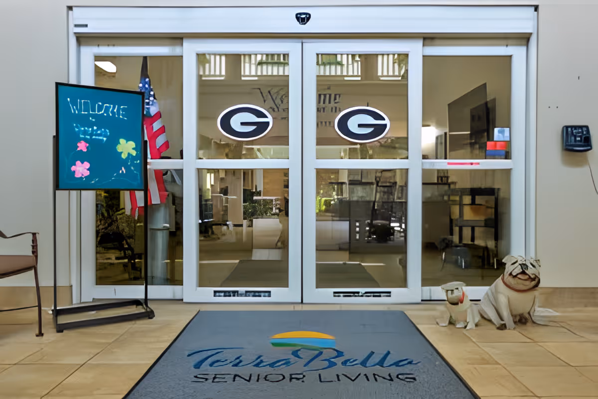 Entrance area of TerraBella Senior Living facility with glass automatic doors featuring two 'G' logos, a welcome sign on a stand with colorful flowers and the word 'WELCOME', an American flag, a chair on the left, two decorative dog statues on the right, and a floor mat with the TerraBella Senior Living logo.