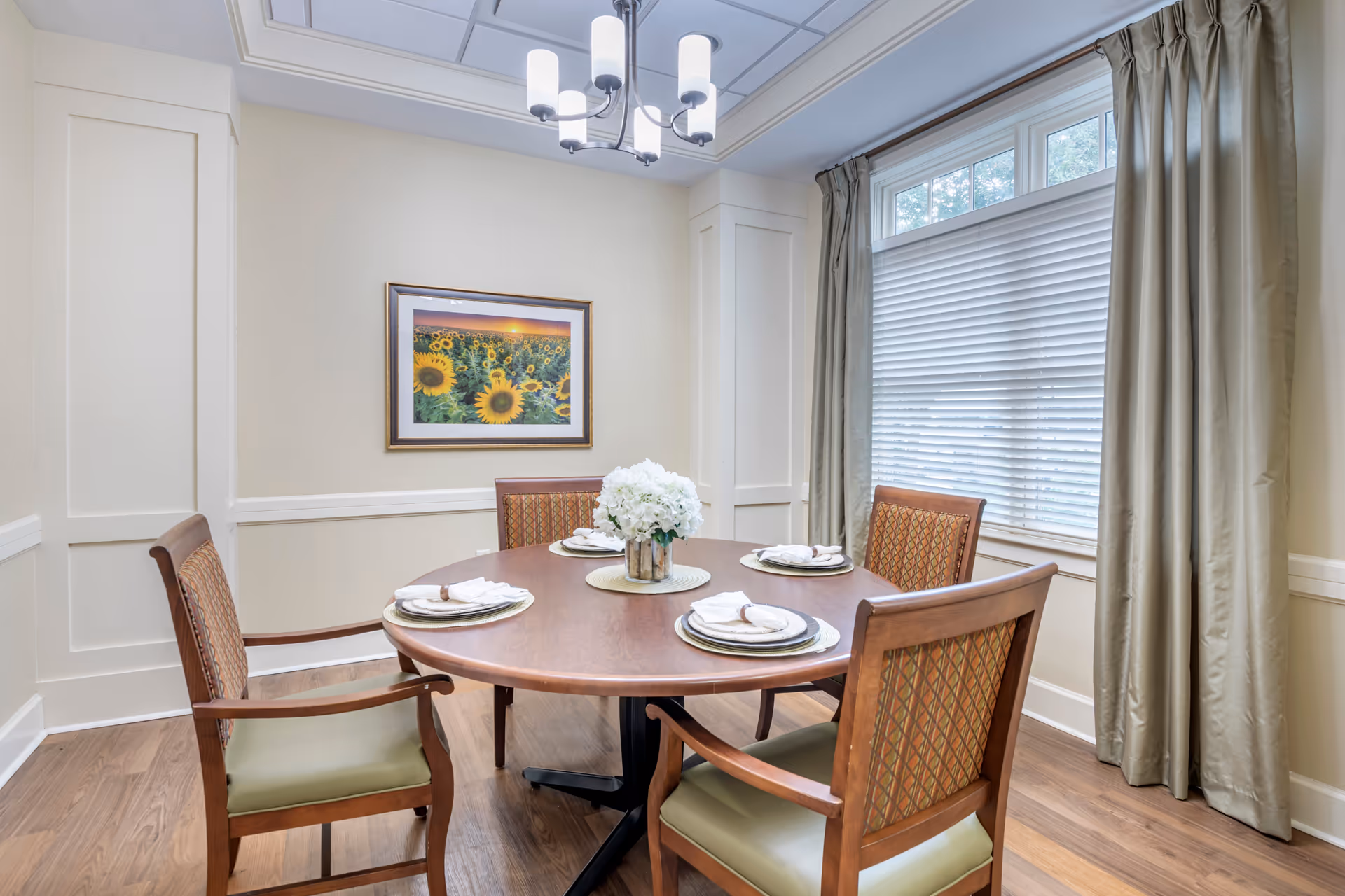 A dining room with a round wooden table set for four, each place setting with a plate and napkin. Four wooden chairs with patterned backs and green cushions surround the table. A vase with white flowers is centered on the table. The room has light-colored walls, a large window with blinds and curtains, a framed picture of sunflowers on the wall, and a modern ceiling light fixture.