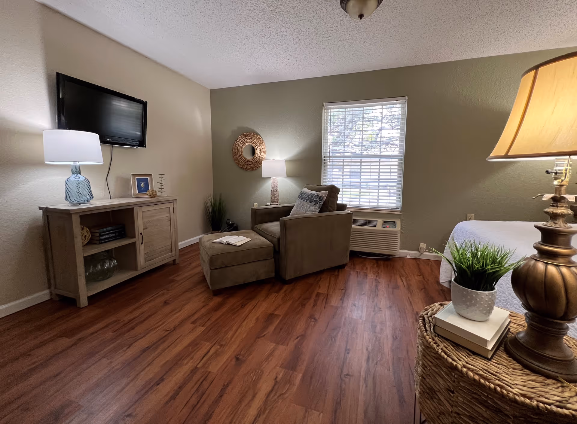 A cozy living area with a brown armchair and matching ottoman placed near a window with blinds. A wooden cabinet with a lamp and decorative items sits against the wall with a flat-screen TV mounted above it. The room has wood flooring, a green accent wall, and a wicker side table with a lamp and a small potted plant.