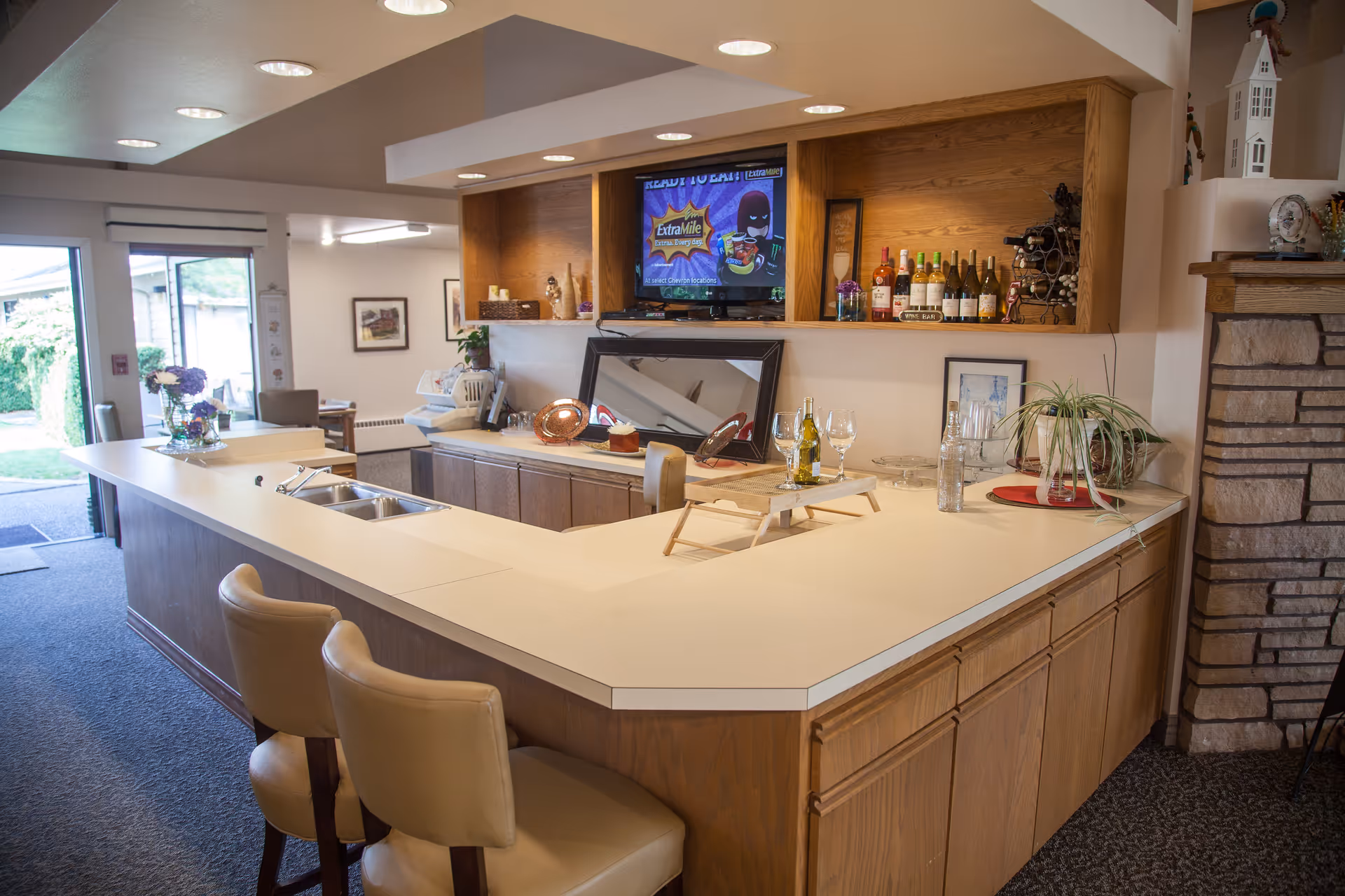 Interior view of a retirement community common area featuring a large L-shaped counter with two beige bar stools. The counter has a built-in sink and is decorated with a small plant, wine bottles, glasses, and a tray. Behind the counter, there are wooden cabinets, a mounted TV, and a stone fireplace to the right. The area is well-lit with recessed ceiling lights and has a carpeted floor. A door to the outside is visible in the background.