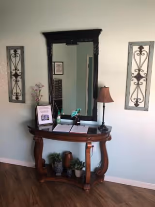 An ornate wooden console table with a large black-framed mirror and decorative items against a pale wall in an interior hallway.